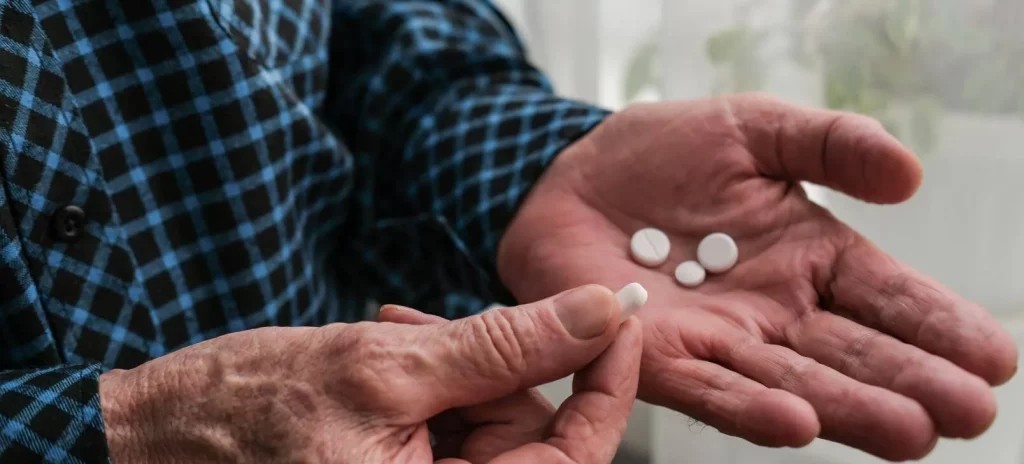 Bejaarde man heeft pillen in zijn linkerhand en met de rechterhand houdt hij een pil vast