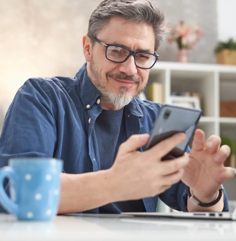 Man van middelbare leeftijd zit aan tafel met een kop koffie en bekijkt glimlachend zijn mobiele telefoon
