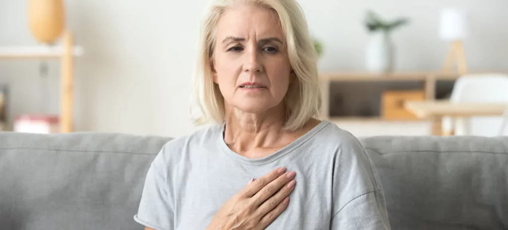 oudere, blonde vrouw zit op de bank en houdt haar hand op haar borst vanwege pijn