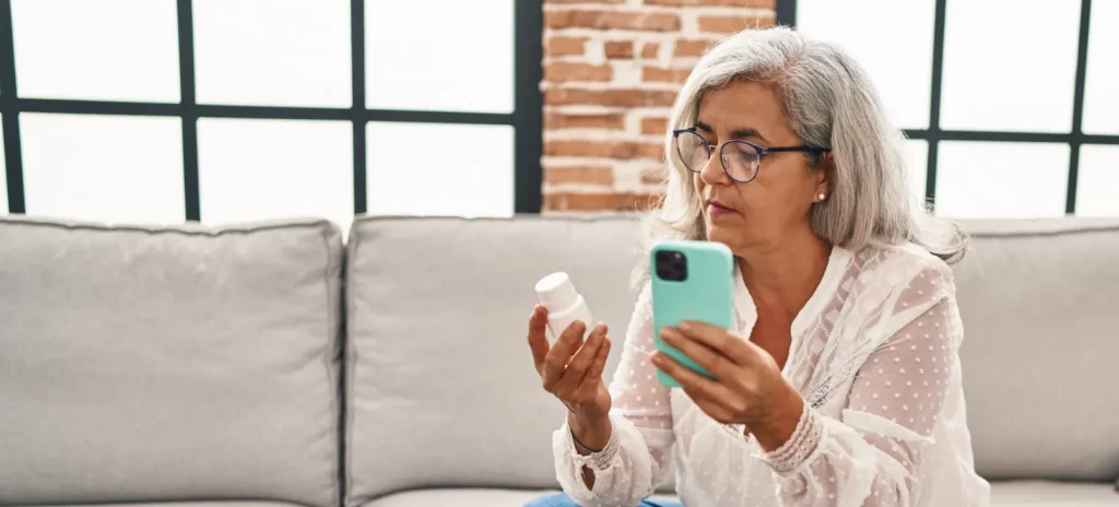 Oudere vrouw zit op de bank en controleert een potje met pillen met haar mobiel in de hand